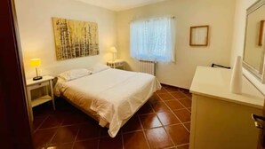 Ein Schlafzimmer mit einem Bett, Nachttischlampen, einem Heizkörper, einem Fenster mit Vorhängen und einem an der Wand hängenden Kunstwerk.
