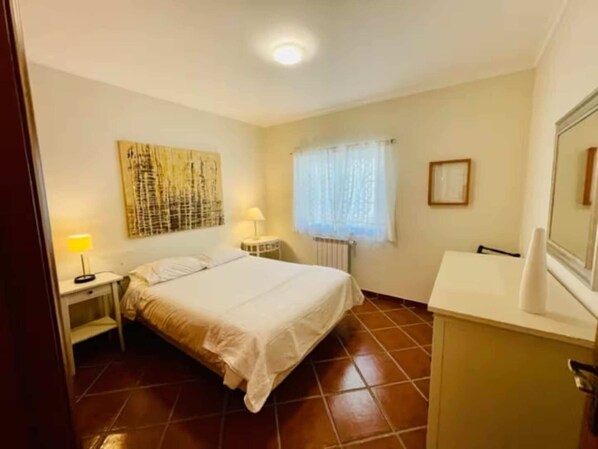 Une chambre à coucher comprenant un lit, des lampes de chevet, un radiateur, une fenêtre avec des rideaux et une œuvre d’art accrochée au mur.
