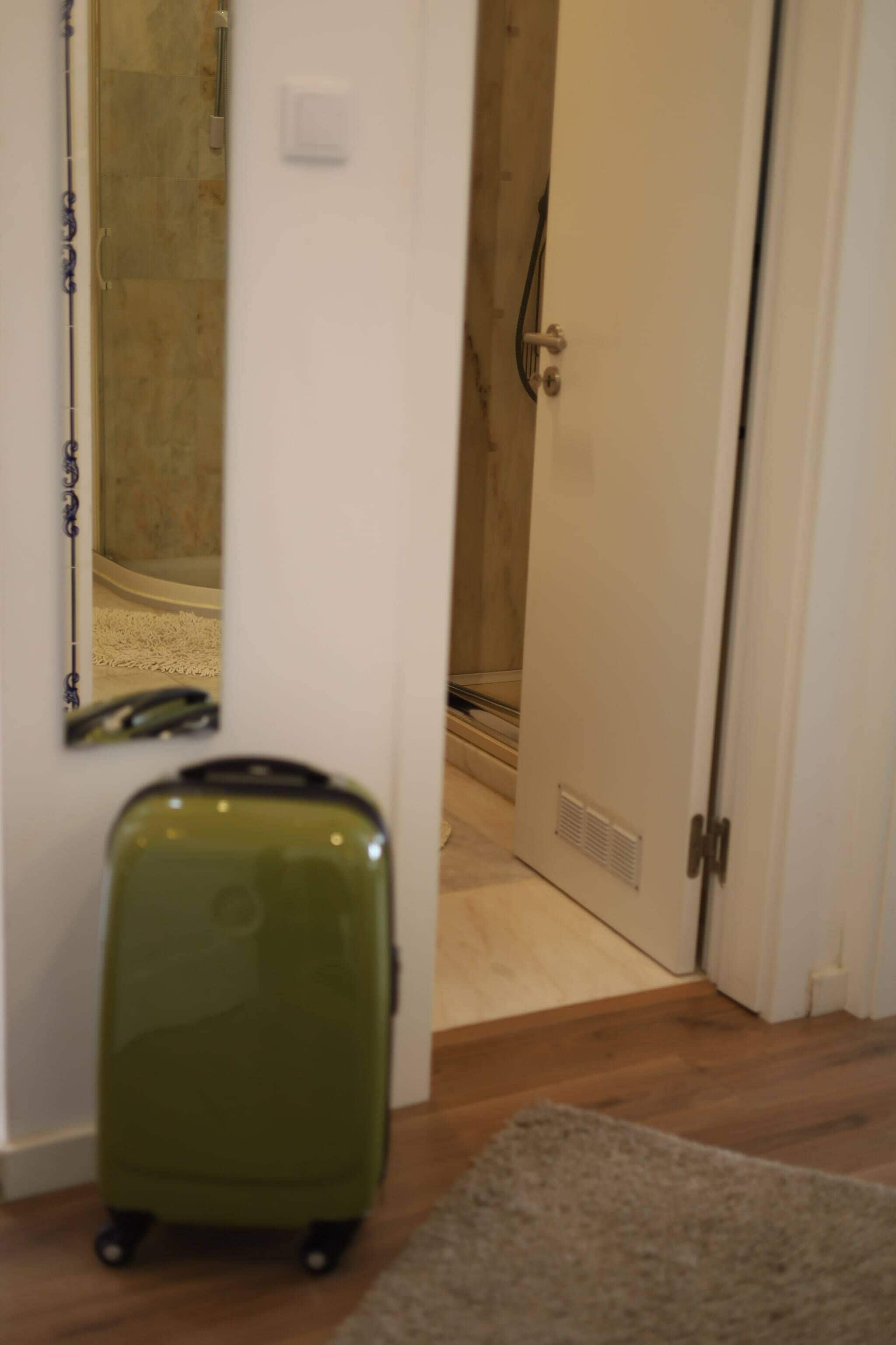 Ein Zimmer mit einem grünen Koffer auf einem Teppich, einer zur Toilette führenden Tür und einem Spiegel an der Wand.