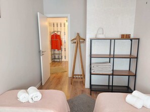 Ein Hotelzimmer mit zwei Betten, einem Kleiderschrank, einem Regal mit Handtüchern und einem zur Toilette führenden Gang.