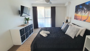 Ein Schlafzimmer mit einem Bett, einem Fernseher, einem Schreibtisch, einer Pflanze und einem Gemälde.