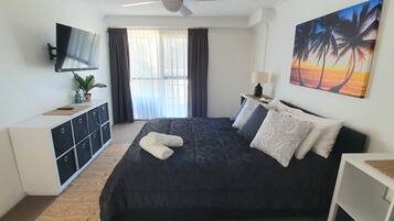 A bedroom with a bed, a TV, a desk, a plant, and a painting.