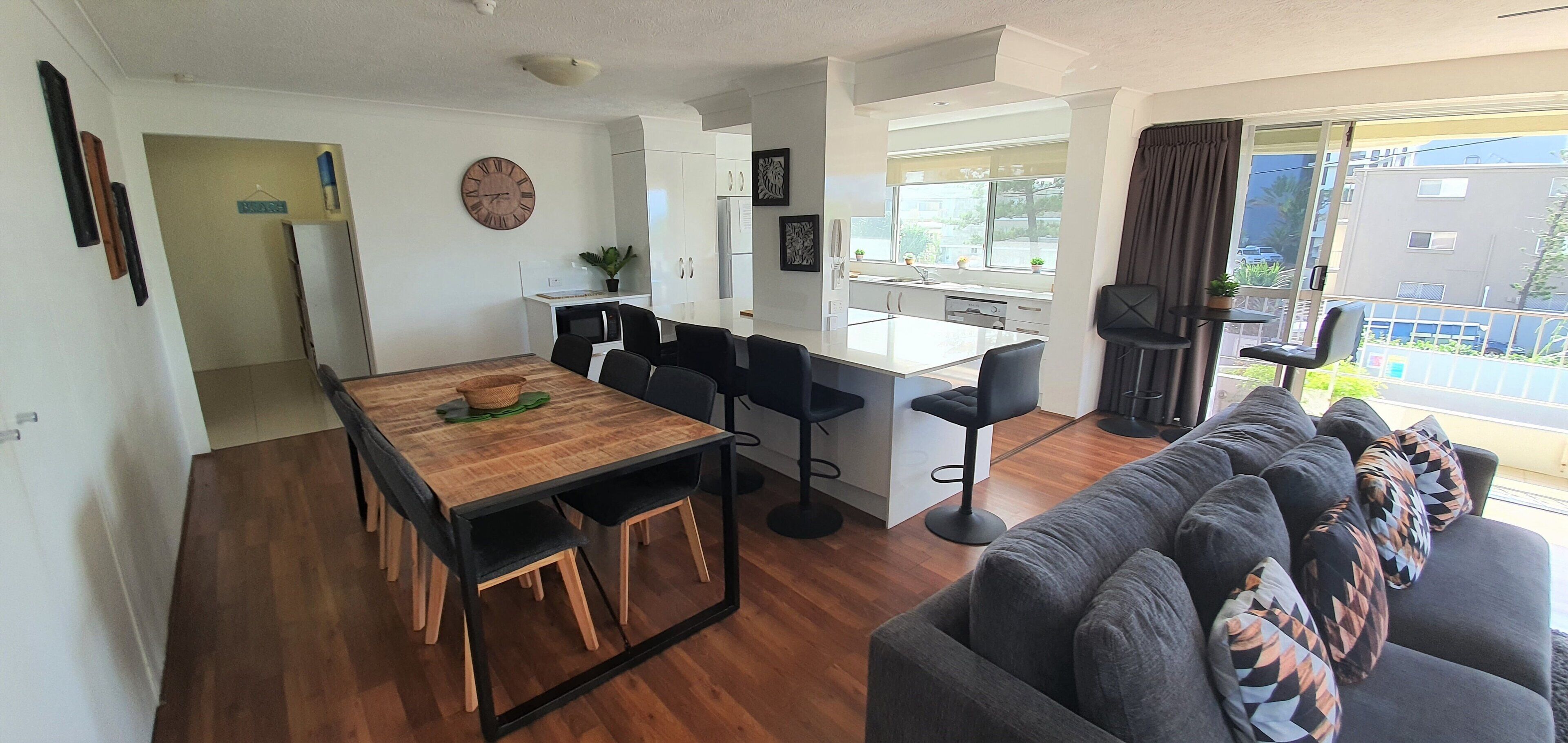A modern living space with a wooden dining table, black bar stools, a grey sofa, and a kitchen area with white cabinets.
