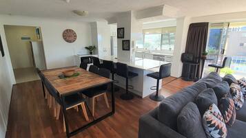 A modern living space with a wooden dining table, black bar stools, a grey sofa, and a kitchen area with white cabinets.