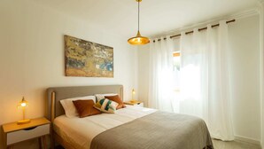 Ett hotellrum med en säng, ett nattduksbord, en lampa, en tavla och ett fönster med gardiner.