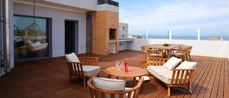 A wooden deck with outdoor furniture, a hanging chair, and a view of the sea.