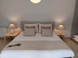 A neatly made bed with two bedside lamps, a radiator, and a small table with a remote control.