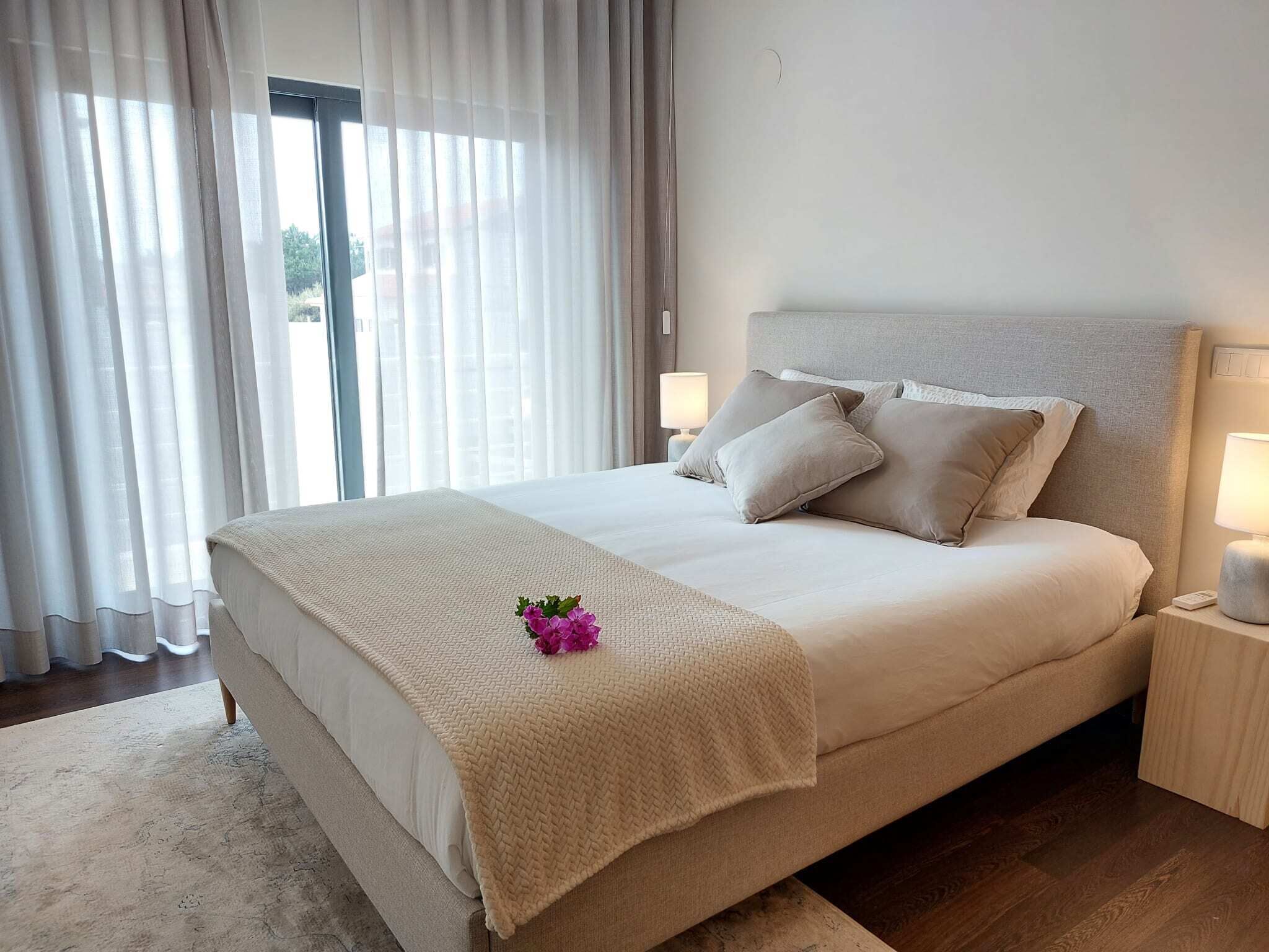 Een hotelkamer met een groot bed, een nachtkastje met een lamp en uitzicht door een raam met gordijnen.