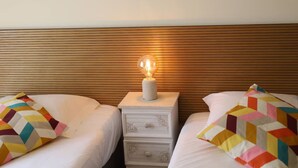 Hotellihuone, jossa on kaksi sänkyä. Jokaisessa sängyssä on värikäs, geometrisesti kuvioitu tyyny, ja sängyn vieressä on yöpöytä, jossa on lamppu. Sängyn päädyssä on puinen koristepaneeli.