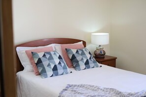 Un lit bien fait, avec une couette blanche, deux coussins à motifs et une lampe de chevet posée sur une table de nuit en bois.