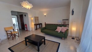 Uma sala de estar com um sofá verde, uma cadeira de madeira, uma mesa de centro preta e um candeeiro suspenso.