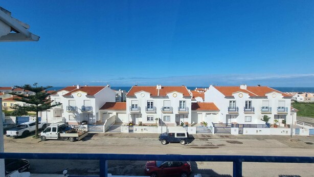 Uma fileira de edifícios brancos com telhados de telha vermelha, um parque de estacionamento com alguns veículos e uma vista desimpedida do mar ao fundo.