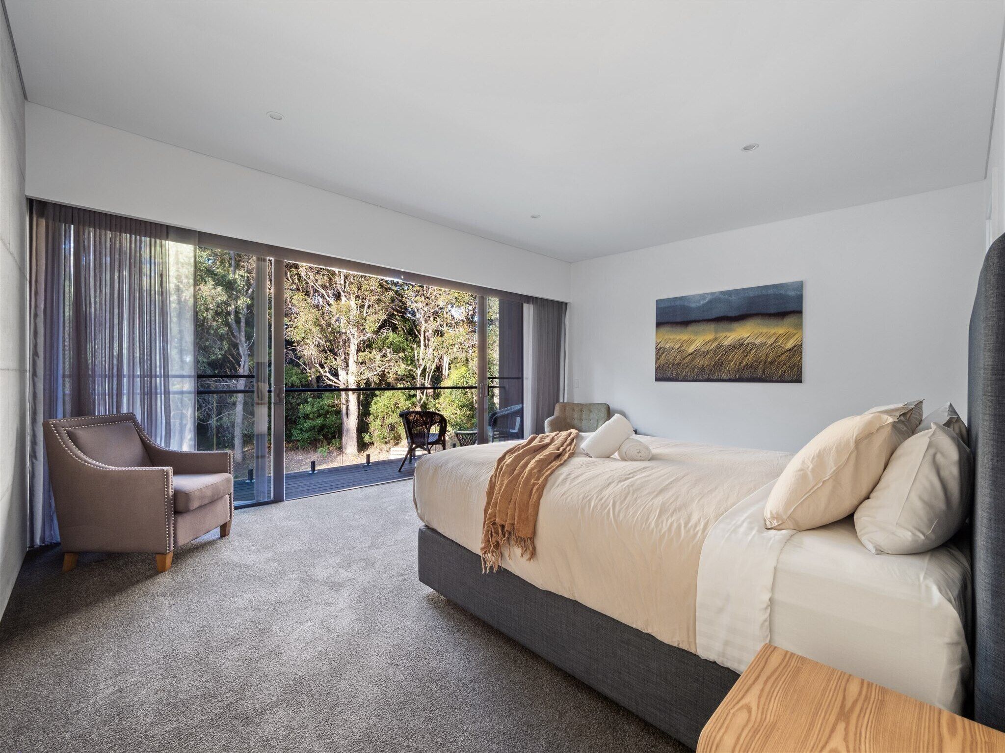 A modern bedroom with a large bed, a chair, and a balcony with a view of trees.