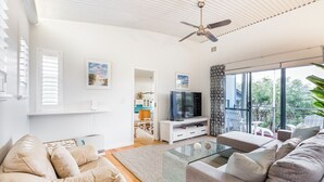 Uma sala de estar moderna com uma mesa de centro de vidro, sofás bege, uma TV de tela plana sobre um suporte branco e um ventilador de teto.