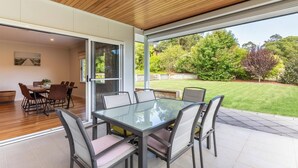 Een moderne eethoek buiten met een glazen tafel en stoelen, grenzend aan een overdekt terras met houten vloer en uitzicht op een verzorgde tuin.