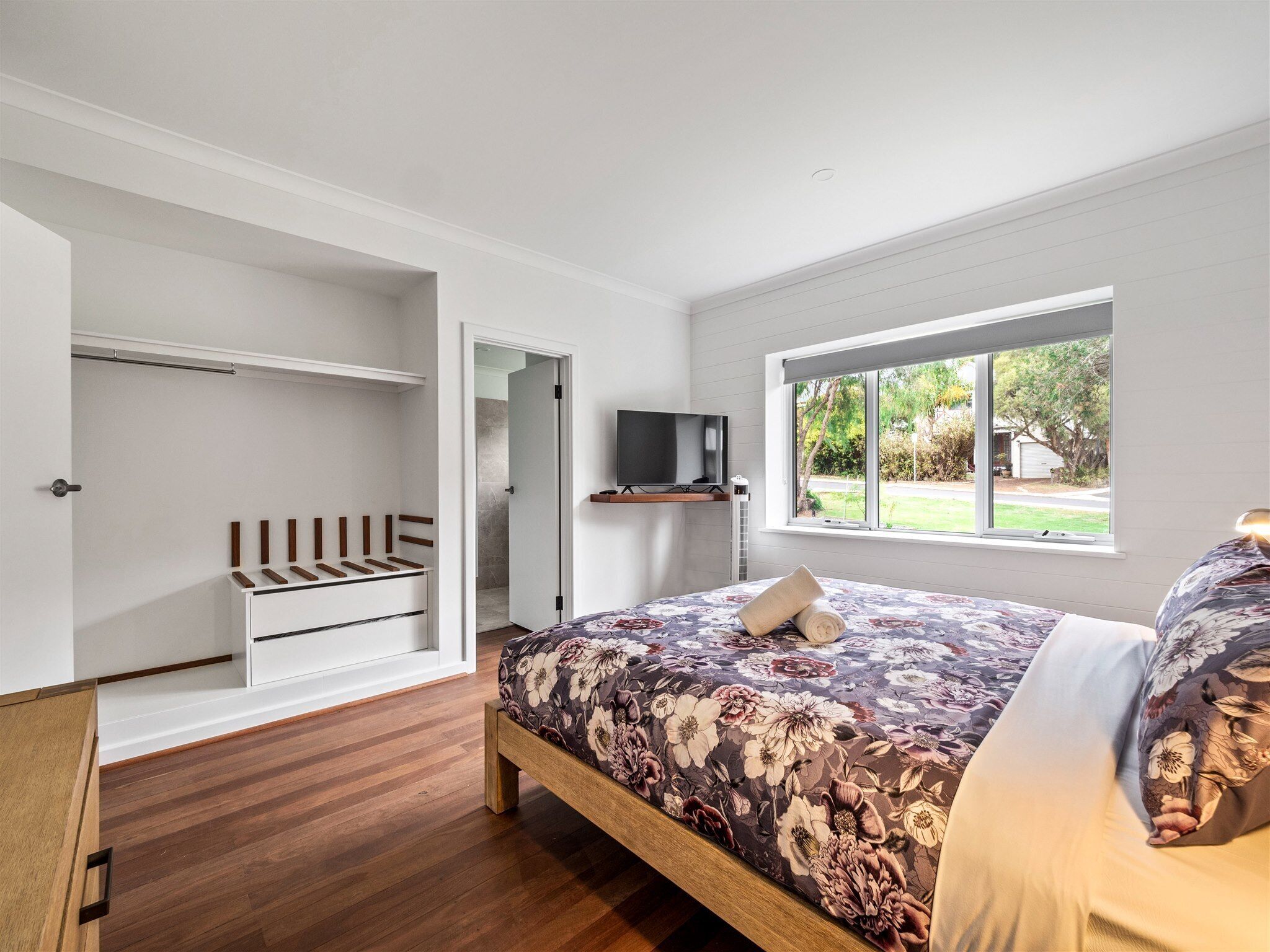 Een slaapkamer met een houten bed, een inbouwkast, een televisie op een plank en een raam met uitzicht op groen.