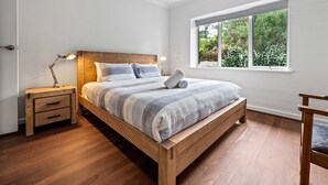 A bedroom with a wooden bed, a nightstand, a desk, and a window with a view of greenery.