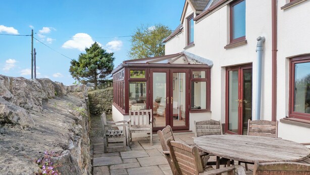 A patio area with a wooden table and chairs, a conservatory with glass doors, and a stone wall. - 5 bedroom accommodation in Horton, near Gower (Horton, near Gower)