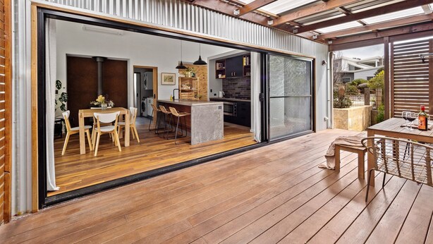 Une vaste terrasse dotée d’un plancher en bois, un coin repas avec une table et des chaises, et un espace cuisine visible à travers une porte-fenêtre coulissante.