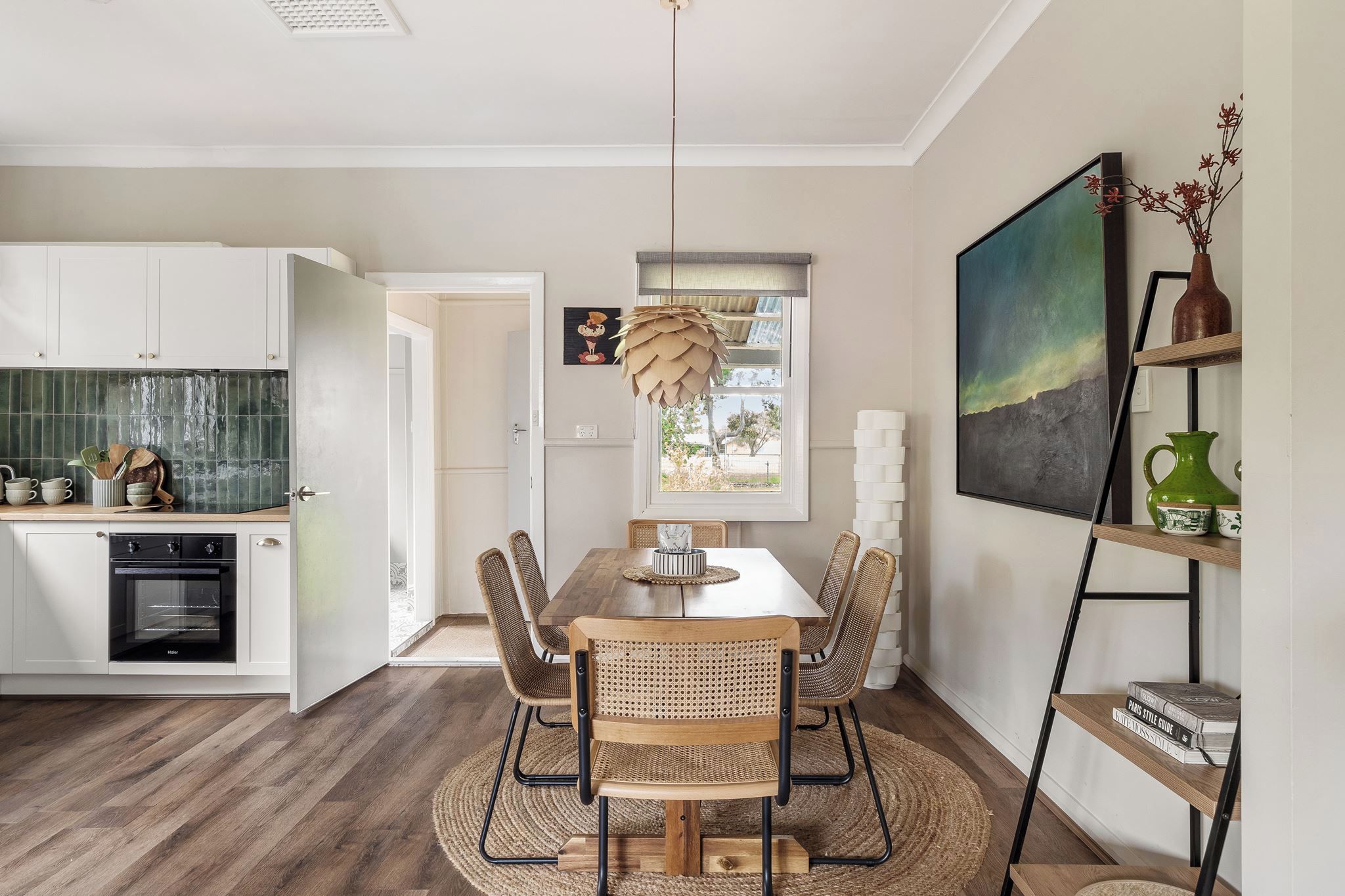 Une cuisine moderne avec des meubles blancs, un four encastré et un coin repas comprenant une table en bois et des chaises en osier.