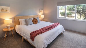 Ein Schlafzimmer mit einem Bett, zwei Nachttischen, zwei Wandlampen, einem Fenster mit Blick auf Bäume und gerahmten Bildern an der Wand.