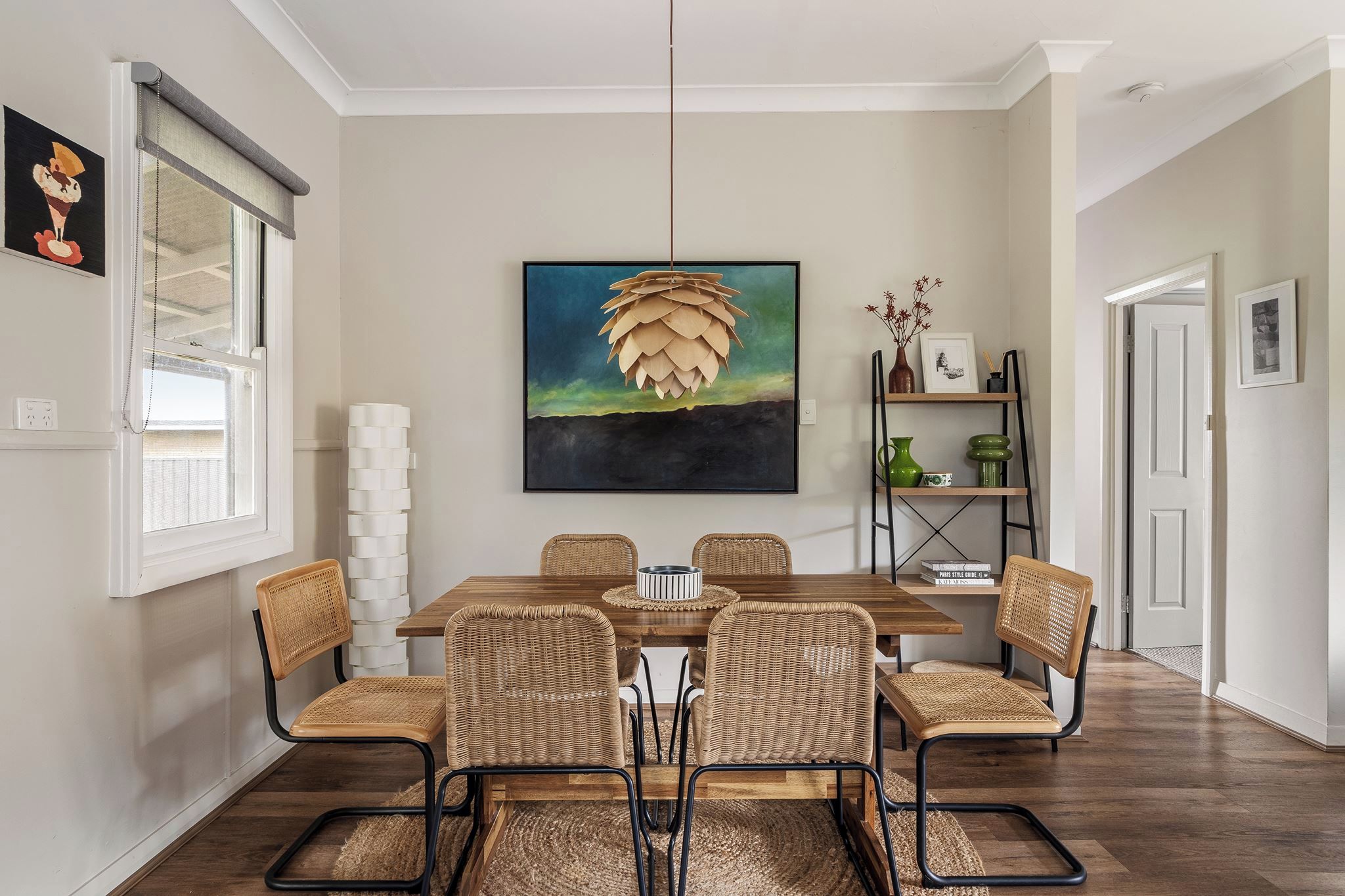 A dining area with a wooden table and wicker chairs, a hanging light fixture, and a painting on the wall.