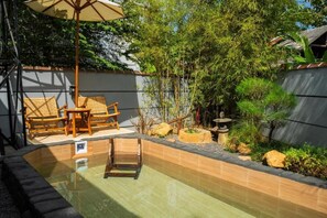 Een zwembad met een houten terras en ligstoelen, omgeven door bamboeplanten en rotsen.