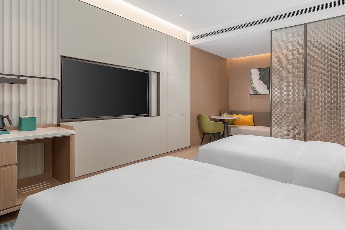 A modern hotel room with two beds, a desk with a coffee maker, a TV, and a dining area with a chair and table.