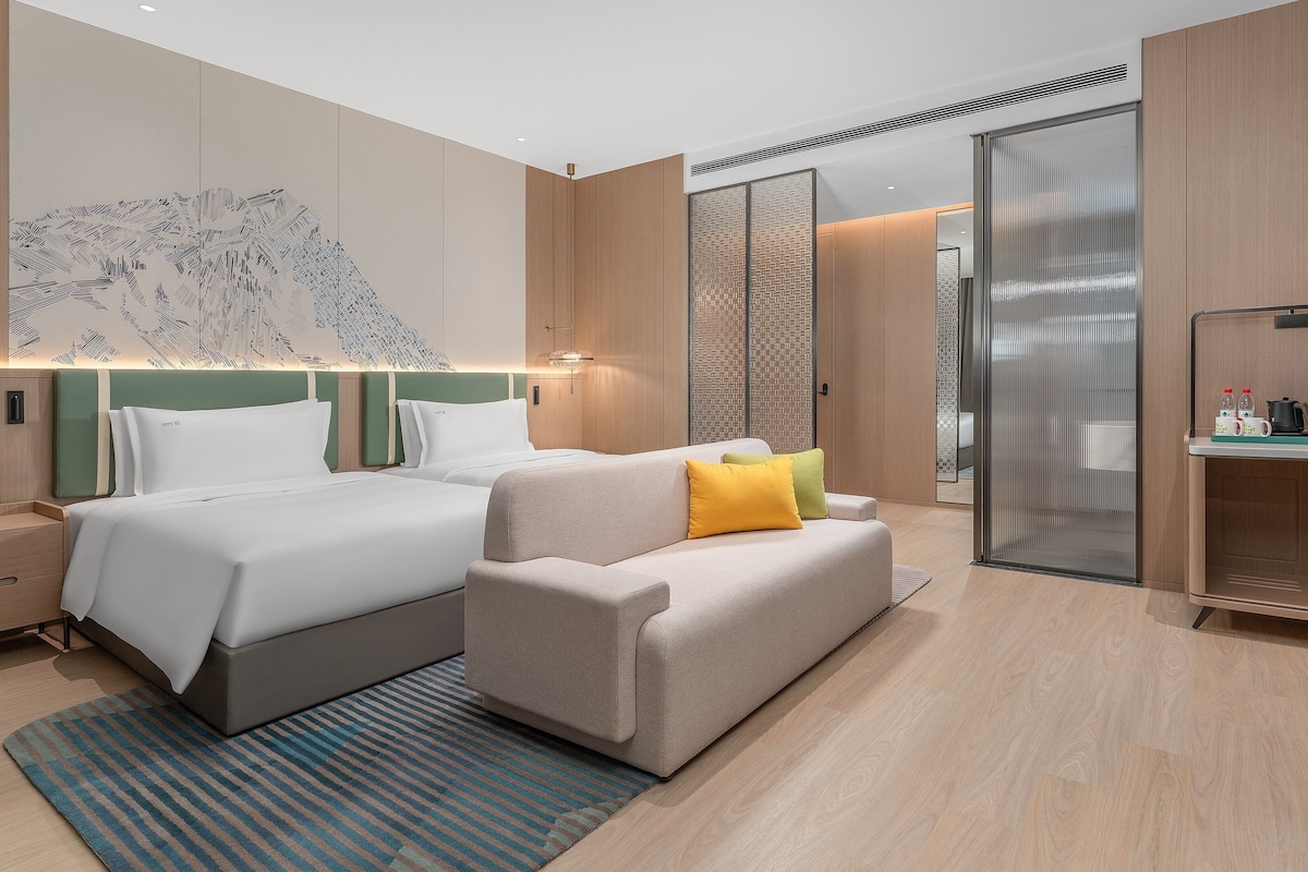 A modern hotel room with two beds, a sofa, a desk, and a large artwork on the wall.
