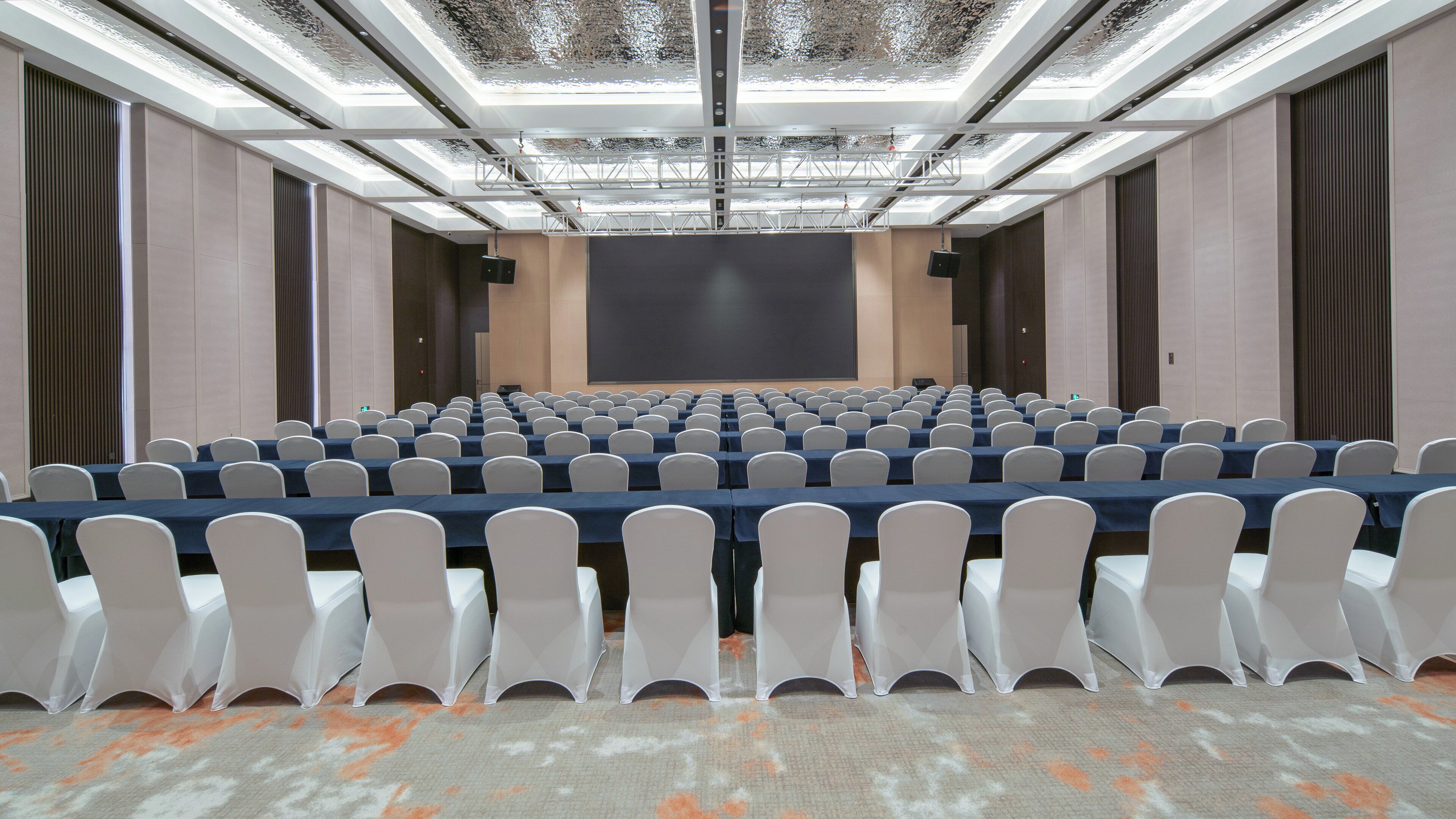 Ein Konferenzraum mit Tischen, Stühlen, einer großen Leinwand und einer hohen Decke mit Oberlichtern.
