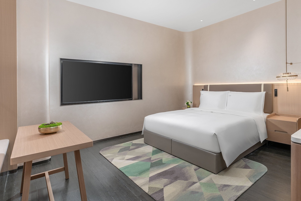 A modern hotel room with a large bed, a bedside table, a small table with a plant, a wall-mounted TV, and a bathroom with a green-tiled shower.