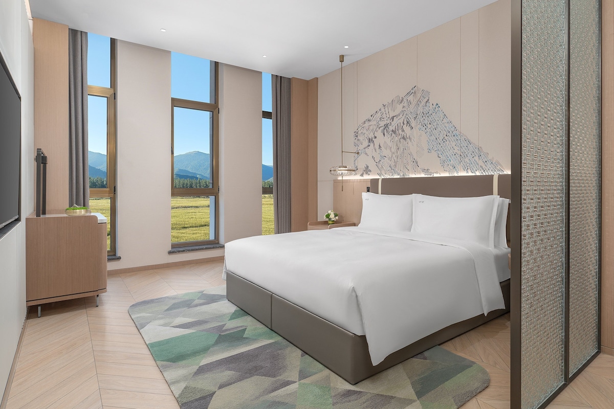 A modern bedroom with a large bed, a view of a mountainous landscape, and a decorative wall piece.