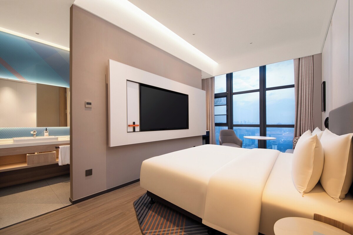 A modern hotel room with a large bed, a TV mounted on the wall, a bathroom with a sink and mirror, and a view of the city through large windows.