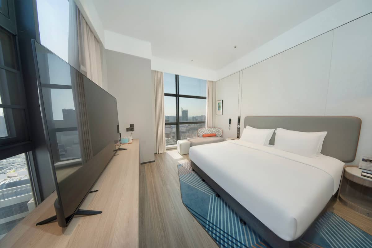 A modern hotel room with a large bed, a flat-screen TV, and a city view.