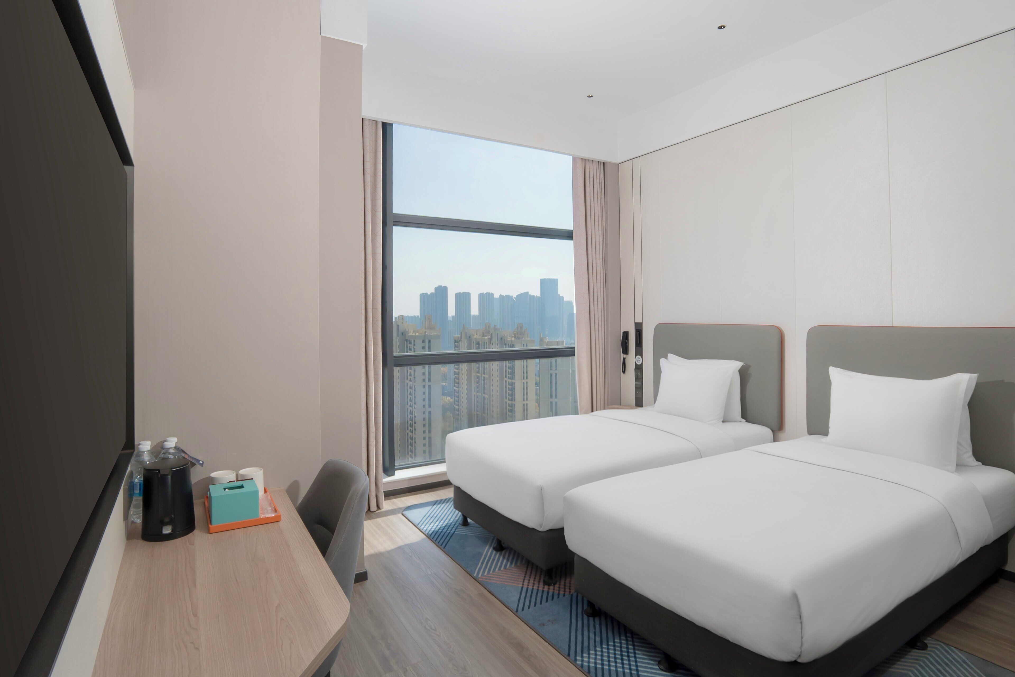 A hotel room with two beds, a desk with a coffee maker, and a large window offering a city view.
