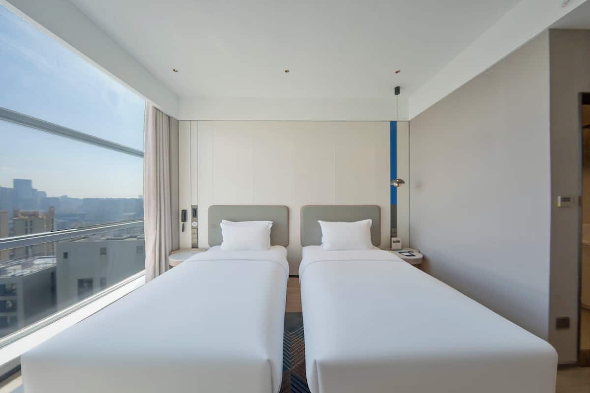 A hotel room with two beds, a large window with a city view, and a door to the right.