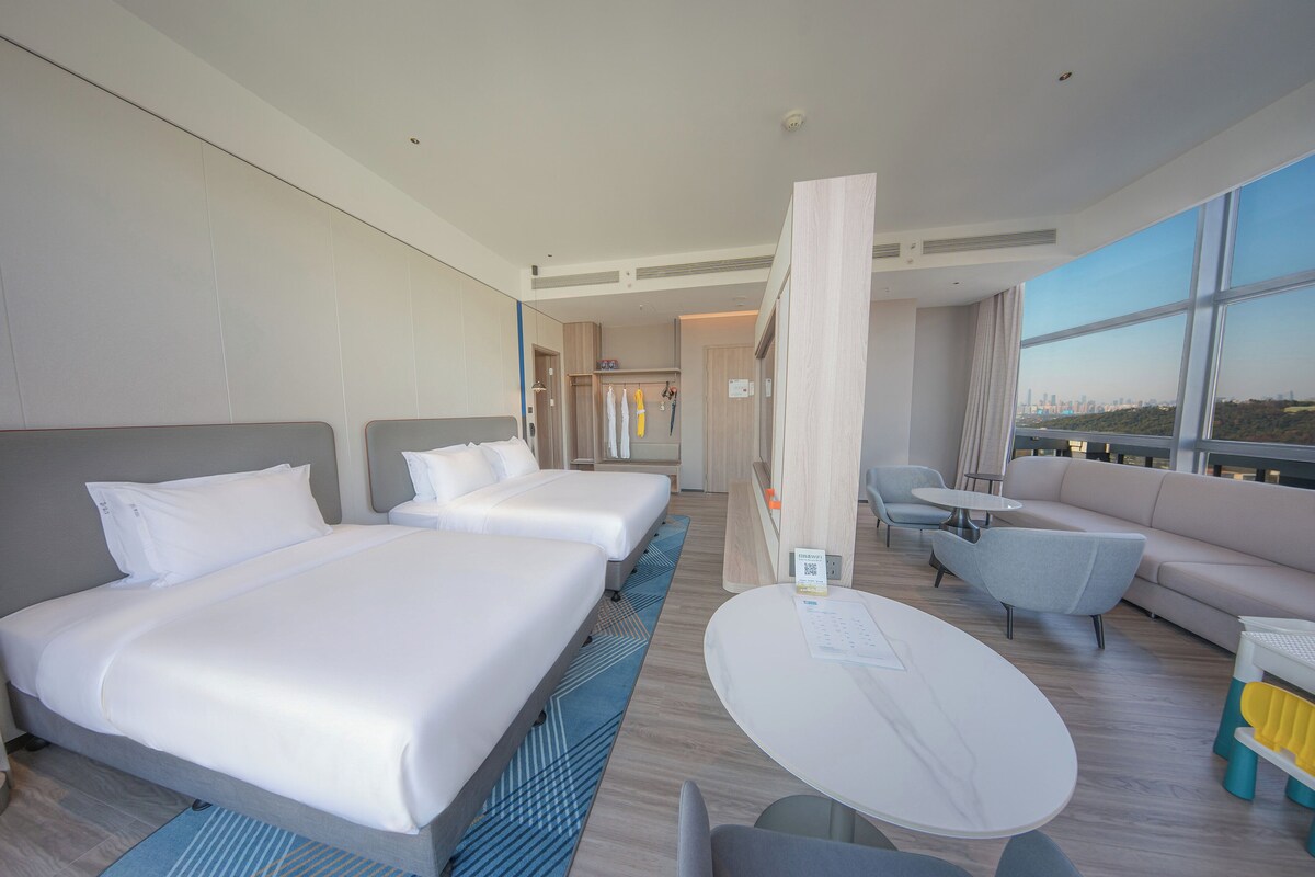 A modern hotel room with two beds, a dining table, a sofa, and a view of the city skyline.