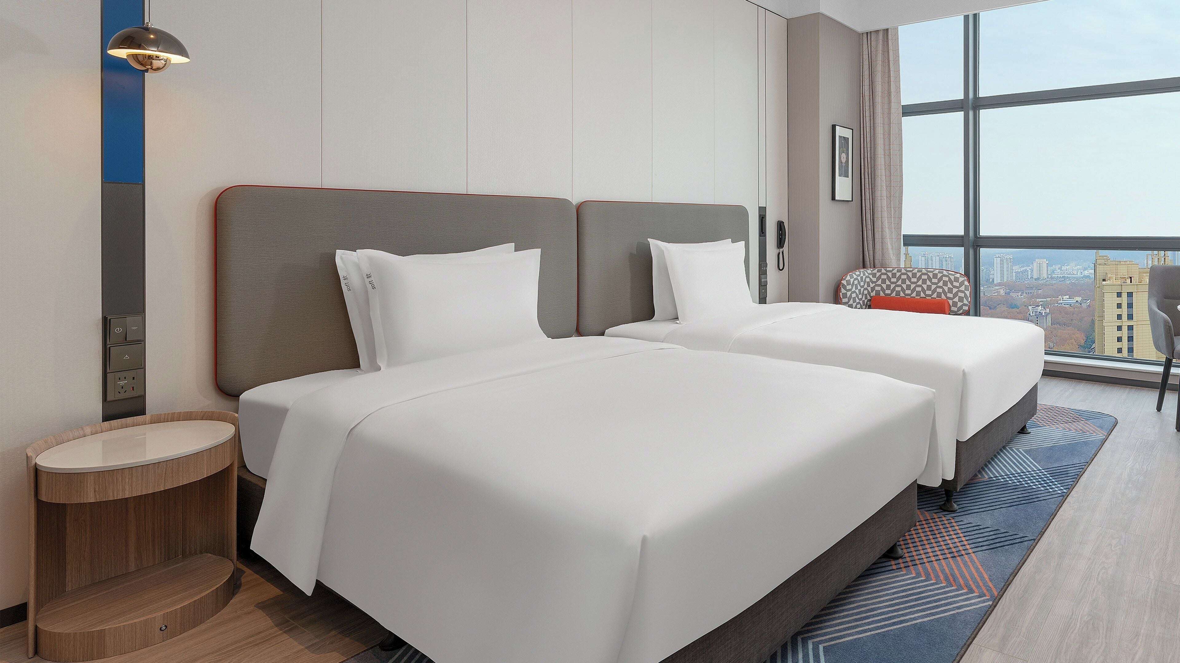 A hotel room with two beds, a small round table, a lamp, and a large window with a city view.
