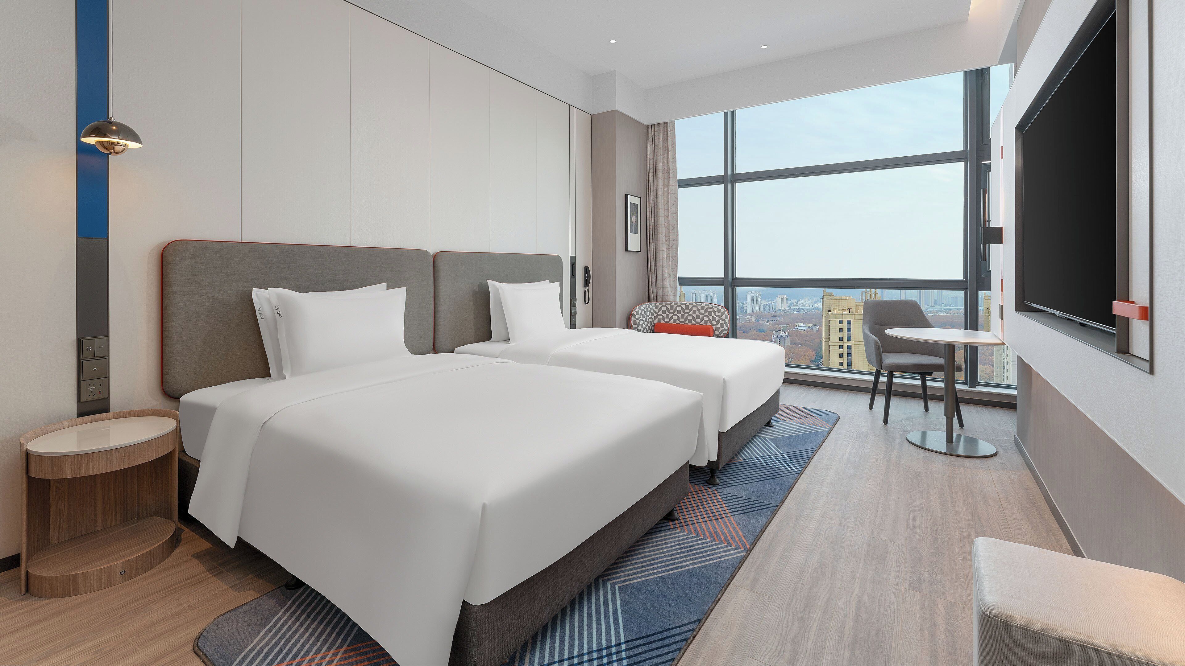 A modern hotel room with two beds, a small table, a chair, and a large window offering a city view.