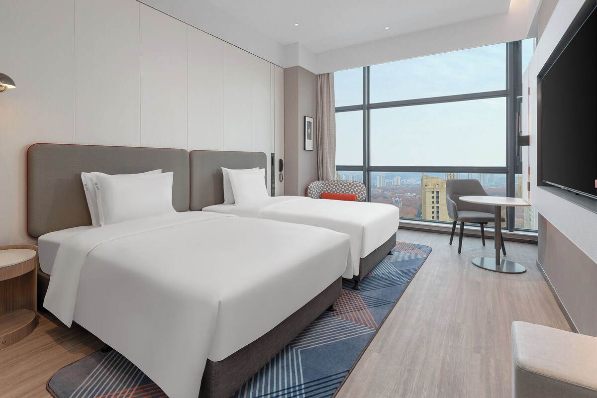 A modern hotel room with two beds, a small table, a chair, and a large window offering a city view.