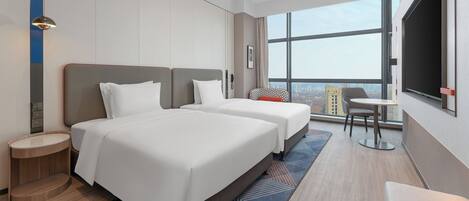 A modern hotel room with two beds, a small table, a chair, and a large window offering a city view.