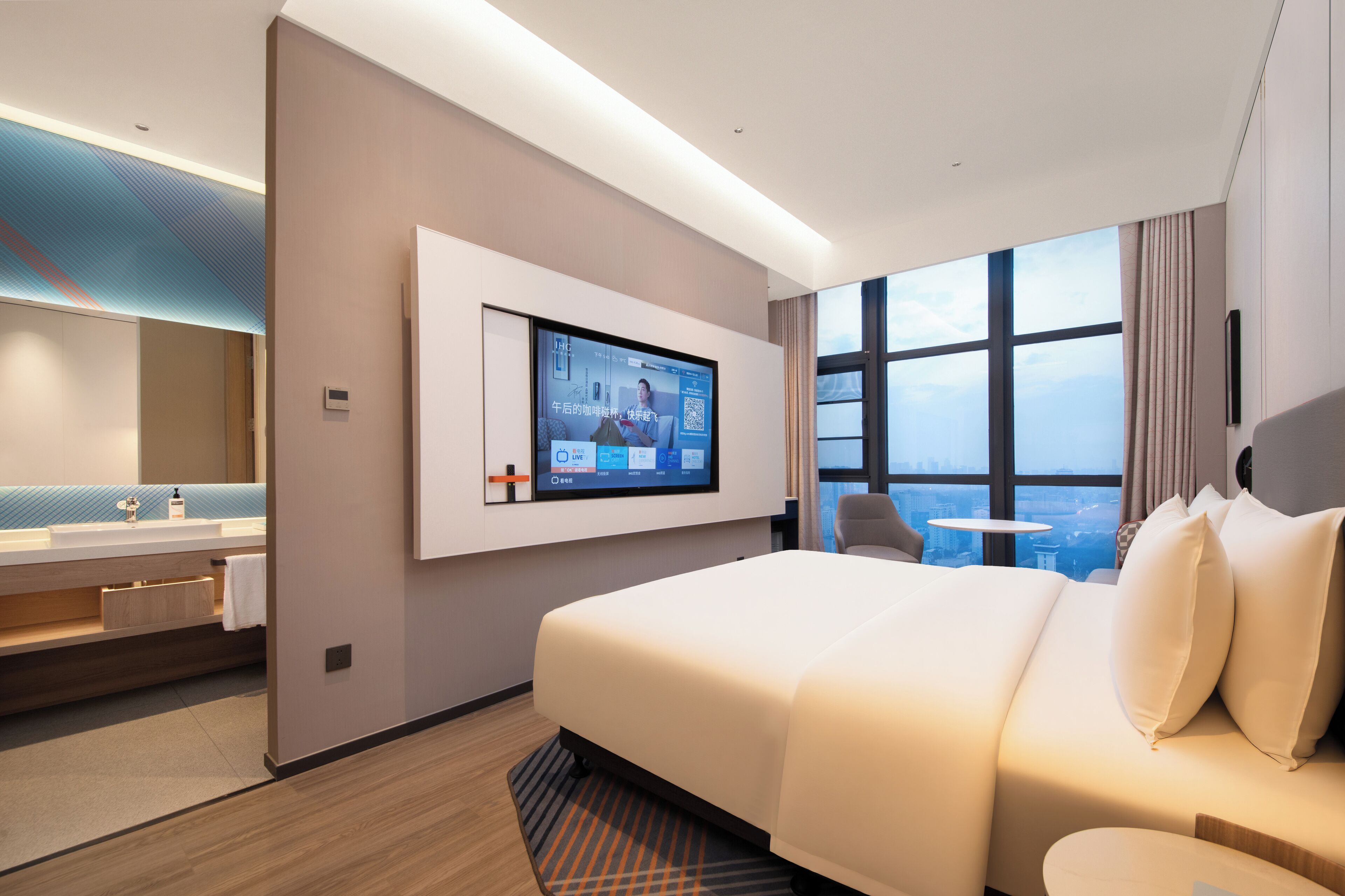 A modern hotel room with a large bed, a TV mounted on the wall, a bathroom with a vanity, and a city view through the window.
