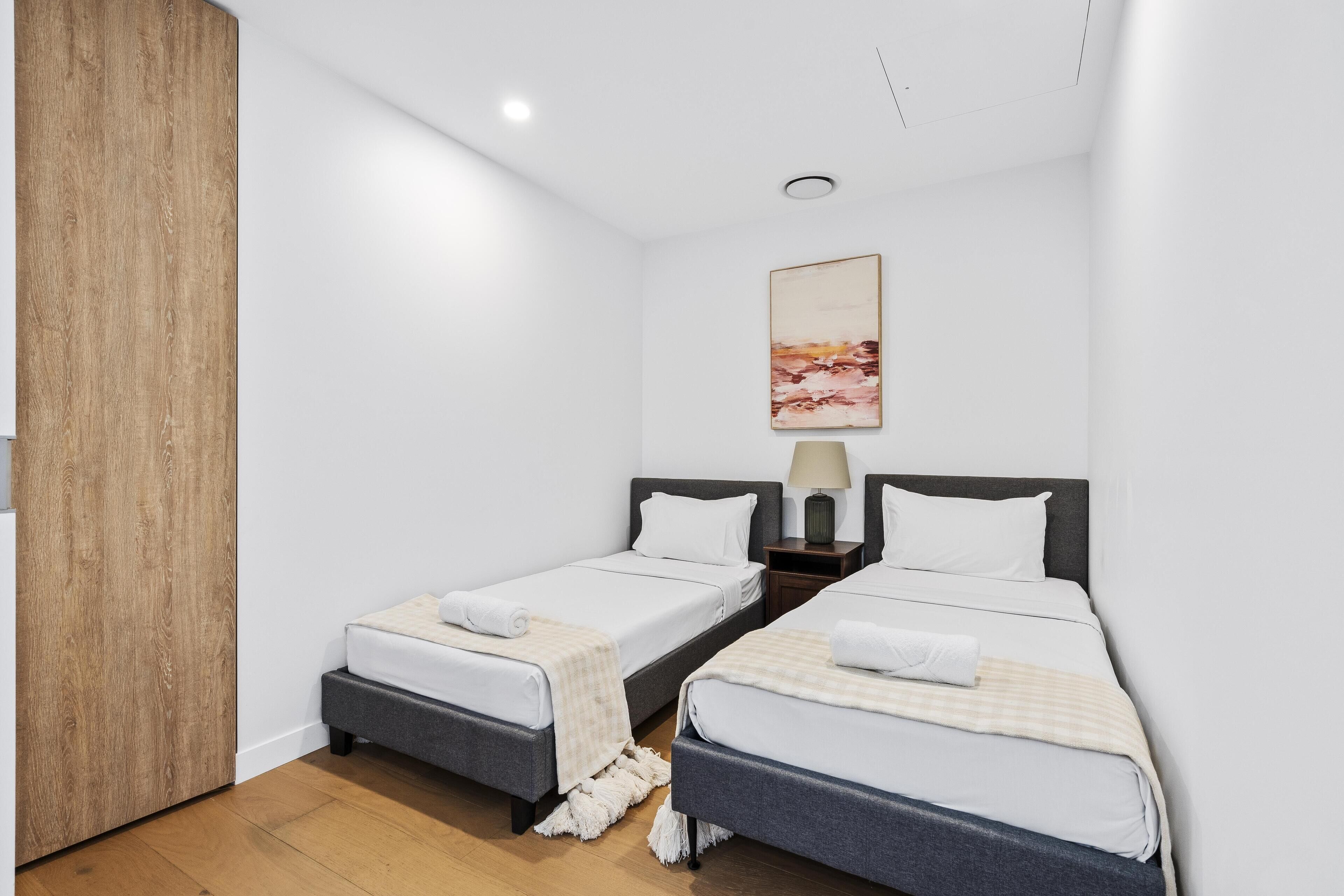 A hotel room with two single beds, a wooden wardrobe, a painting on the wall, and a small nightstand with a lamp.