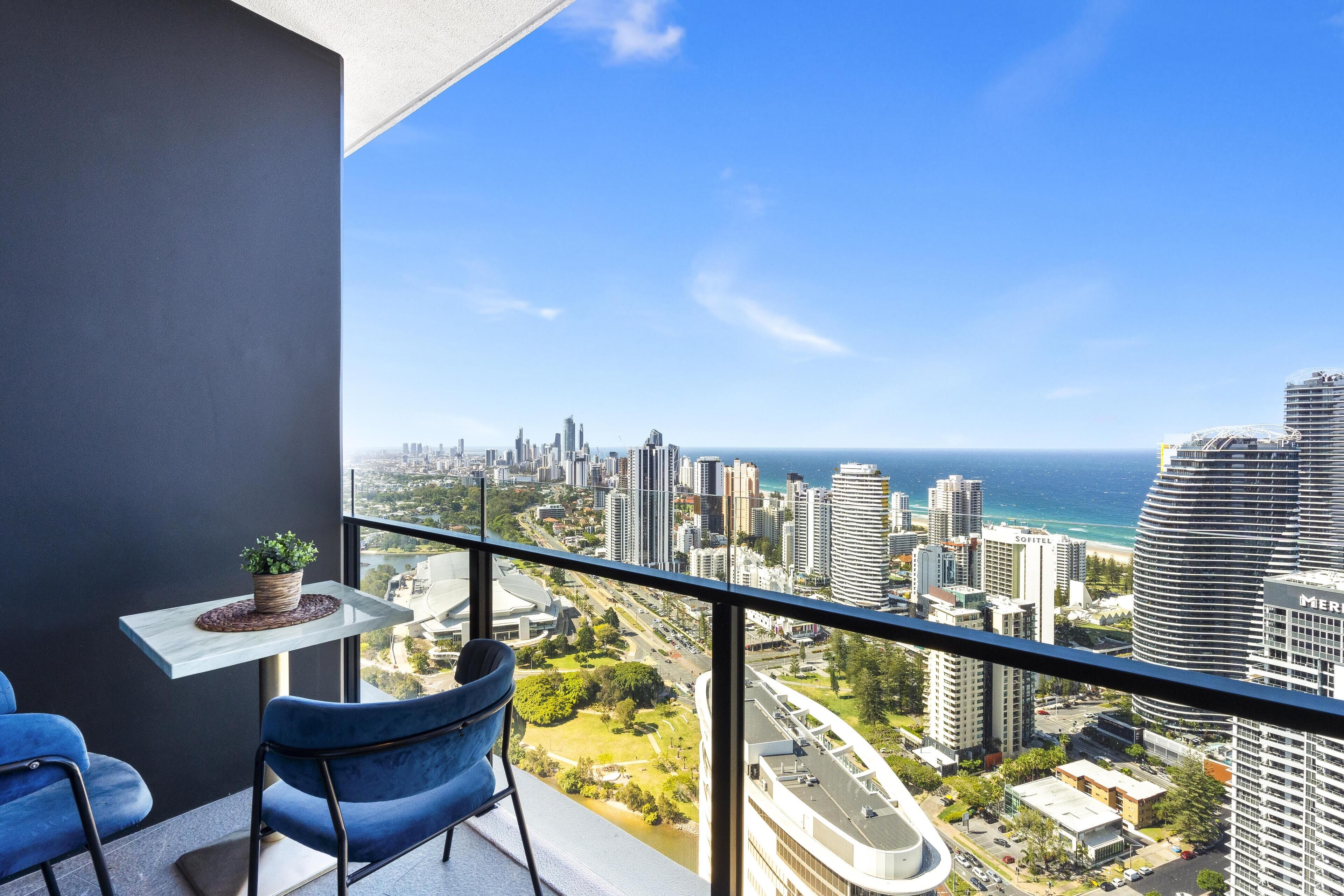 Ein Balkon mit Blick auf die Stadt, ein Tisch mit einer Pflanze und zwei blaue Stühle.