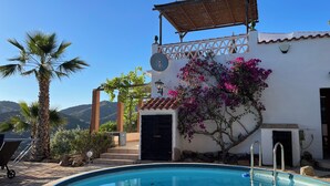 A swimming pool with a clear blue water, surrounded by a white building with a tiled roof, a pergola, and a view of mountains in the background.
