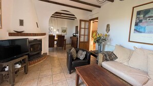 A living room with a fireplace, a sofa, a coffee table, and a TV. There is a dining area in the background with a table and chairs.
