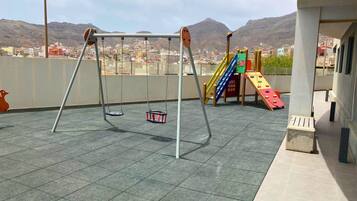 A playground with swings and a slide on a rooftop terrace with a cityscape view.