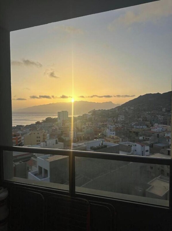 A view from a window overlooking a cityscape at sunset with buildings and a distant mountain range. - Sunset in Mindelo (Mindelo)