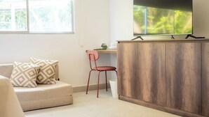A modern living room with a flat-screen TV on a wooden cabinet, a beige sofa with patterned cushions, a red chair, and a window with a view of trees. - Gardin home (Cali)