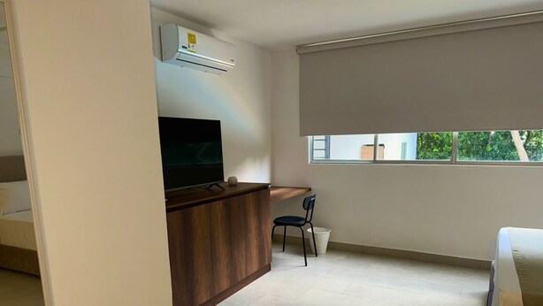 Ein modernes Zimmer mit Schreibtisch aus Holz, einem Sessel, einem Fernseher und einem Fenster mit Blick auf Grünflächen.